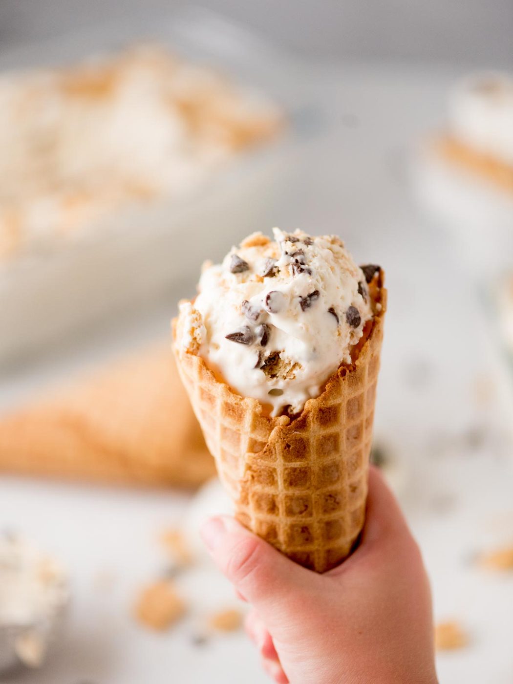 A hand holding a waffle cone filled with ice cream containing chocolate chunks and cookie pieces, with another empty cone and blurred ice cream in the background.