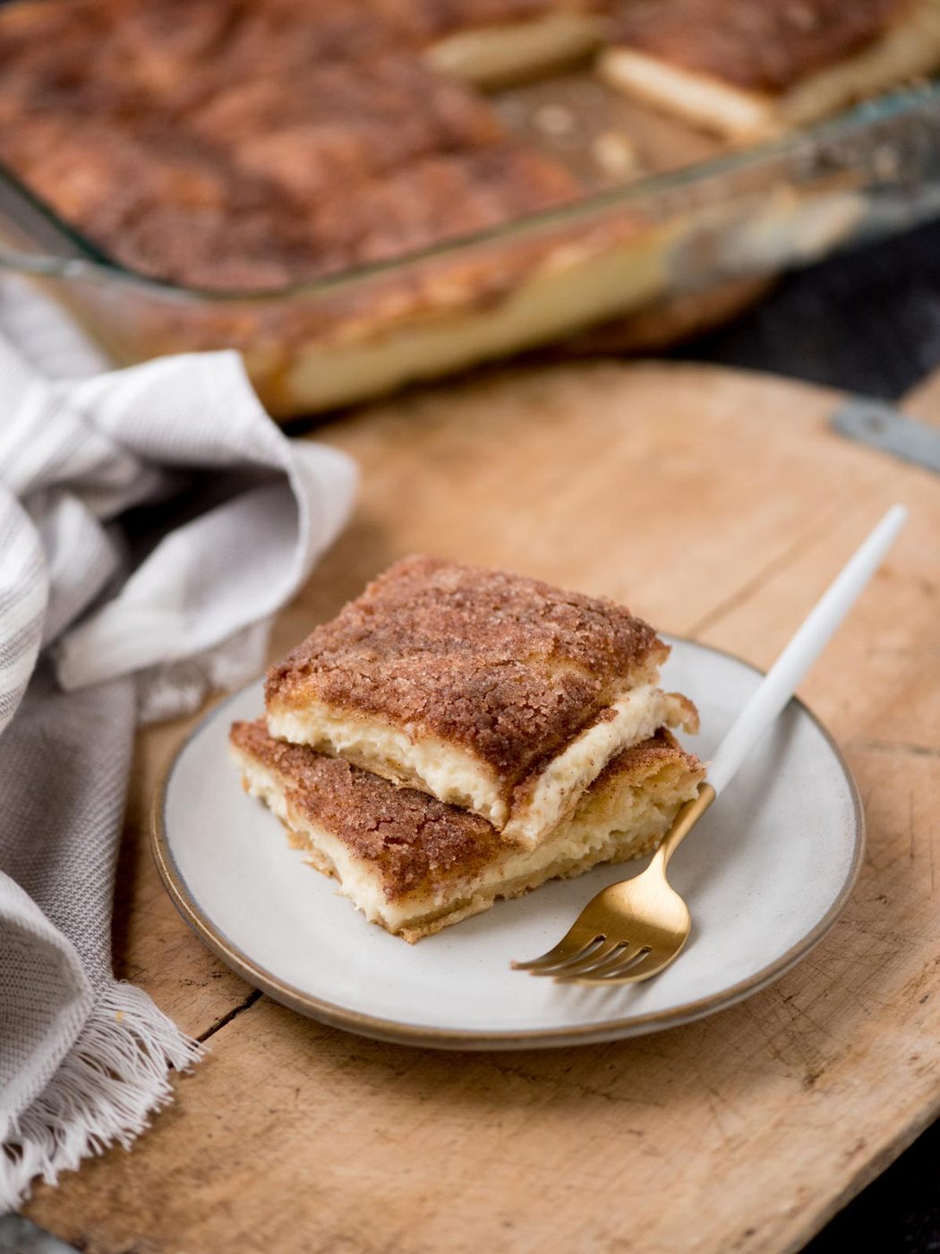 A white plate with two pieces of layered sopapilla cheesecake dessert topped with cinnamon and sugar. A gold fork rests on the plate. In the background, a glass baking dish holds more of the dessert. A striped cloth lies beside the dish on a wooden surface.