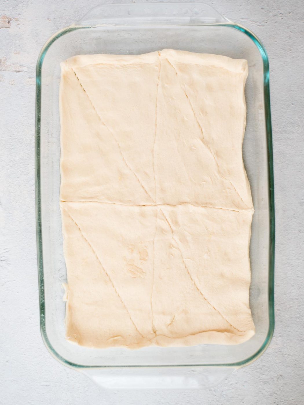 Glass pan containing an unbaked sheet of crescent dough laid flat, covering the bottom evenly. The dough has visible seams and is set on a light gray surface.