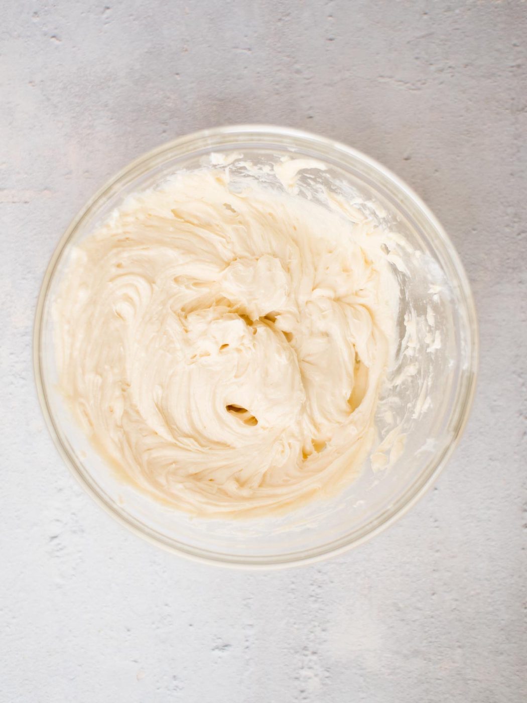 A glass bowl filled with creamy, whipped vanilla frosting sits on a light gray textured surface. The frosting is smooth and swirled, indicating it has been freshly mixed.
