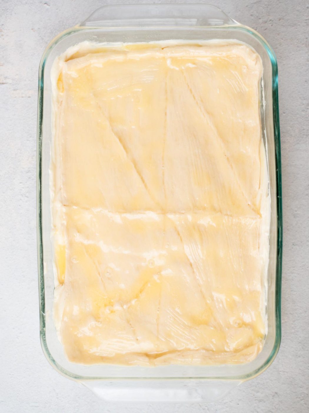 A glass baking dish filled with unbaked dough, scored with diagonal lines. The dough is golden and smooth, ready for baking. The background is a light gray surface.