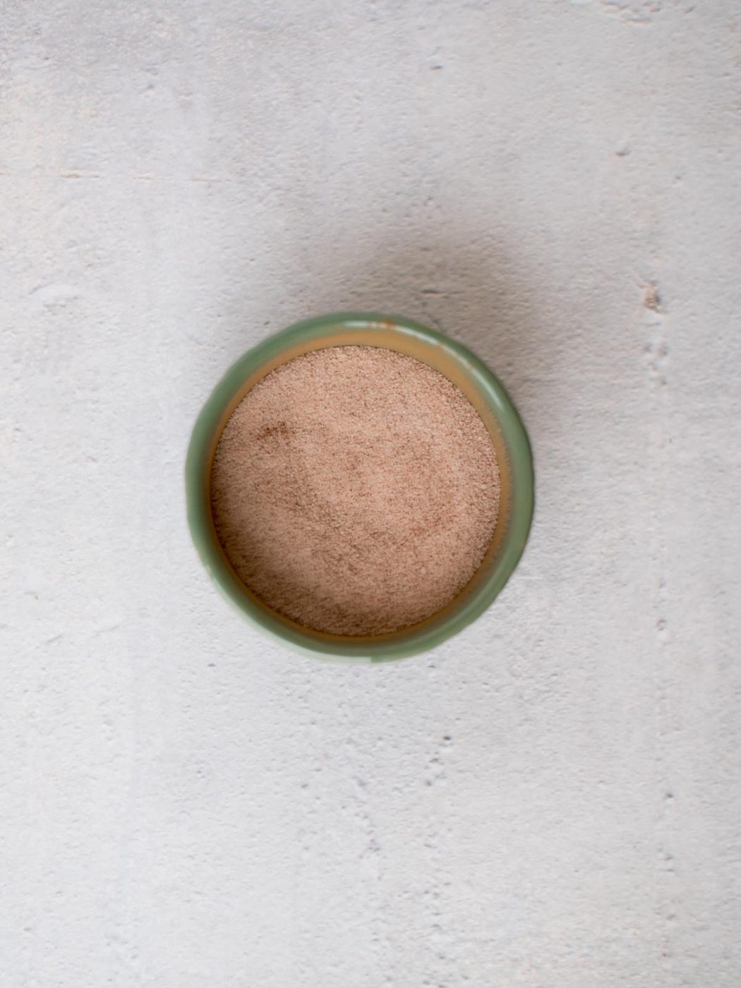 A small, round green bowl filled with a fine cinnamon sugar placed on a textured light gray surface.