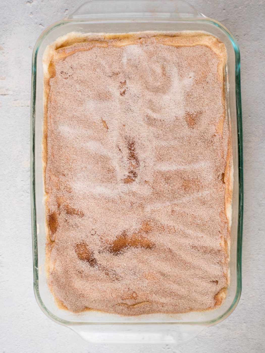 A glass baking dish containing a sopapilla cheesecake dessert sprinkled with a layer of cinnamon sugar, creating a brown and white speckled appearance on the surface.