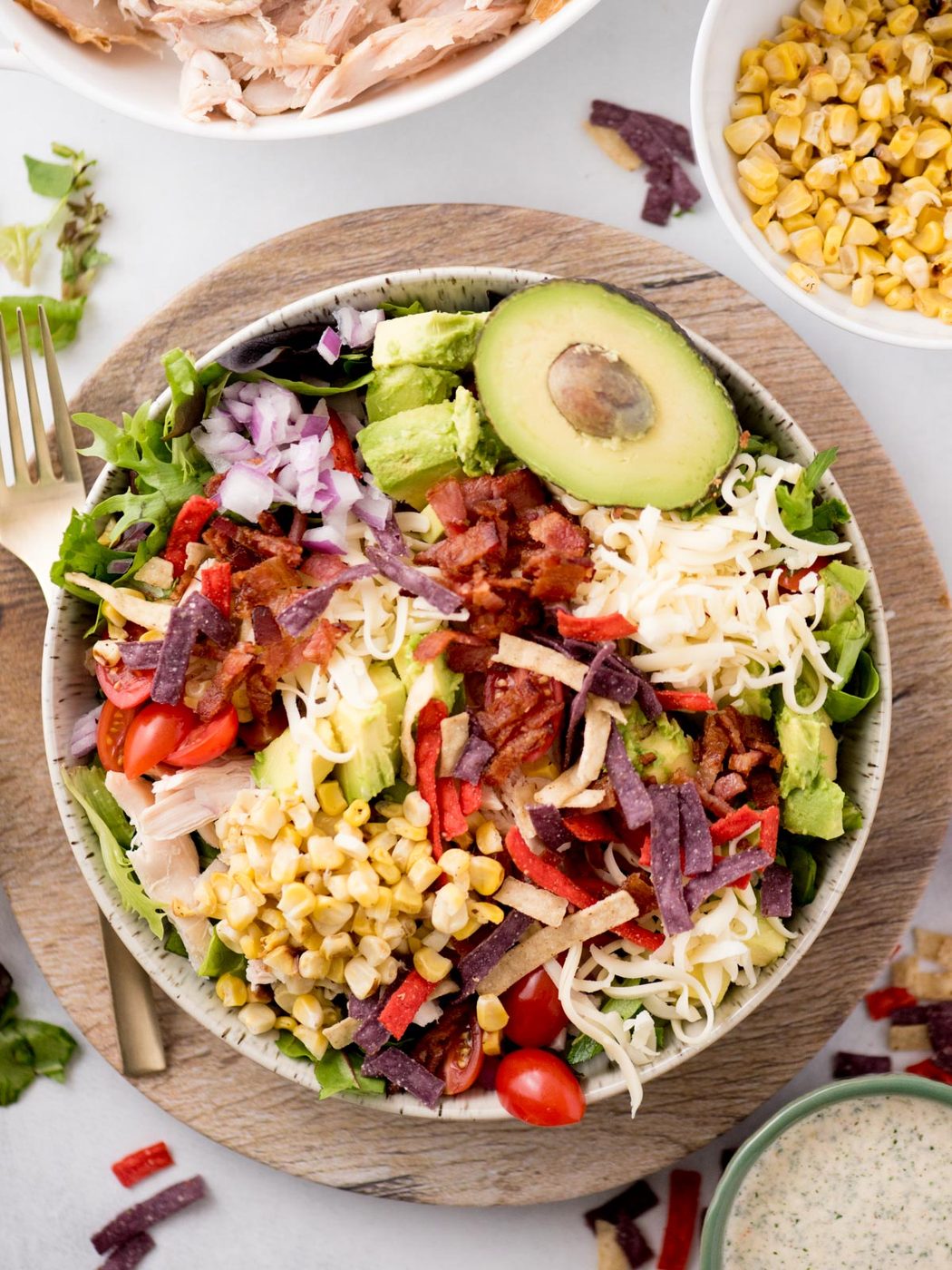 A colorful salad in a bowl with lettuce, sliced avocado, corn, shredded cheese, cherry tomatoes, red onions, bacon bits, and multicolored tortilla strips. A fork lies beside it, with ingredients like corn and dressing in small bowls nearby.