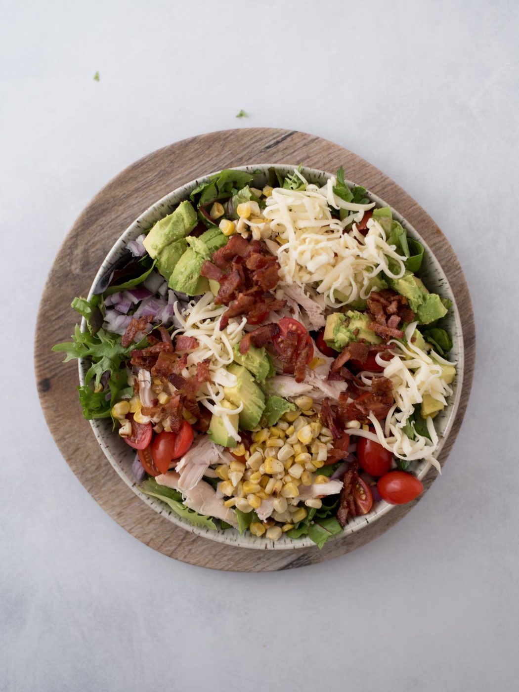 A bowl of salad on a wooden plate, containing shredded cheese, diced avocado, corn, bacon bits, cherry tomatoes, lettuce, and red onion slices.