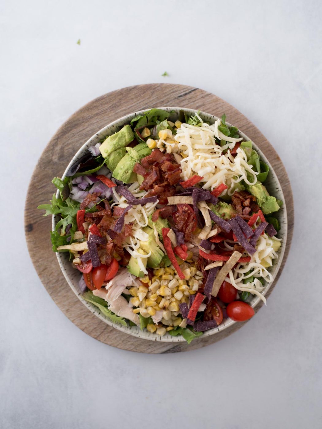 A vibrant salad in a bowl on a wooden plate. It features sliced avocado, shredded cheese, cherry tomatoes, red onions, corn, bacon bits, grilled chicken, and tortilla strips over a bed of greens.