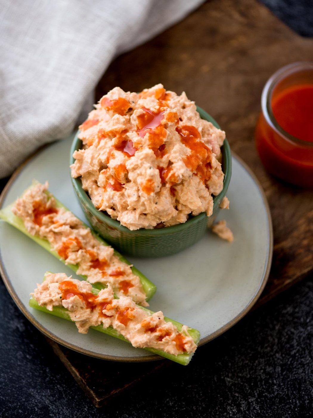 A bowl of spicy buffalo tuna salad on a plate, topped with hot sauce. Next to it are two celery sticks filled with the same dip, drizzled with hot sauce. A cloth and a small jar of spicy buffalo sauce is in the background.