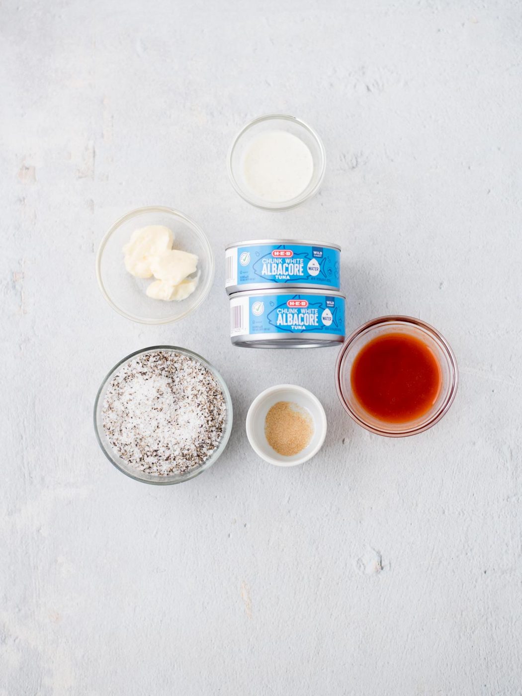 Ingredients arranged on a light surface: two cans labeled Albacore, a bowl of white sauce, a bowl of spicy buffalo sauce, a bowl of salt and pepper, and two small bowls containing liquid and powdered ingredients. Well-balanced for crafting a bold tuna salad with an unexpected kick.