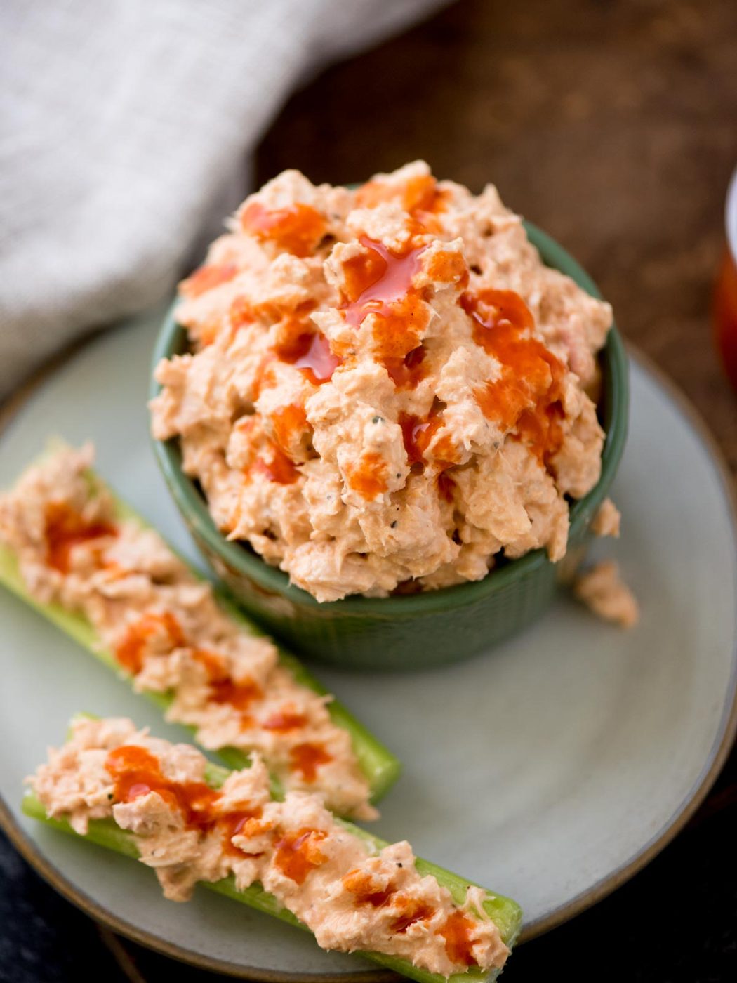 A green bowl brimming with spicy buffalo tuna dip, topped with hot sauce. On a light blue plate beside it, two celery sticks hold the same creamy mixture, drizzled with hot sauce. A light cloth graces the background.