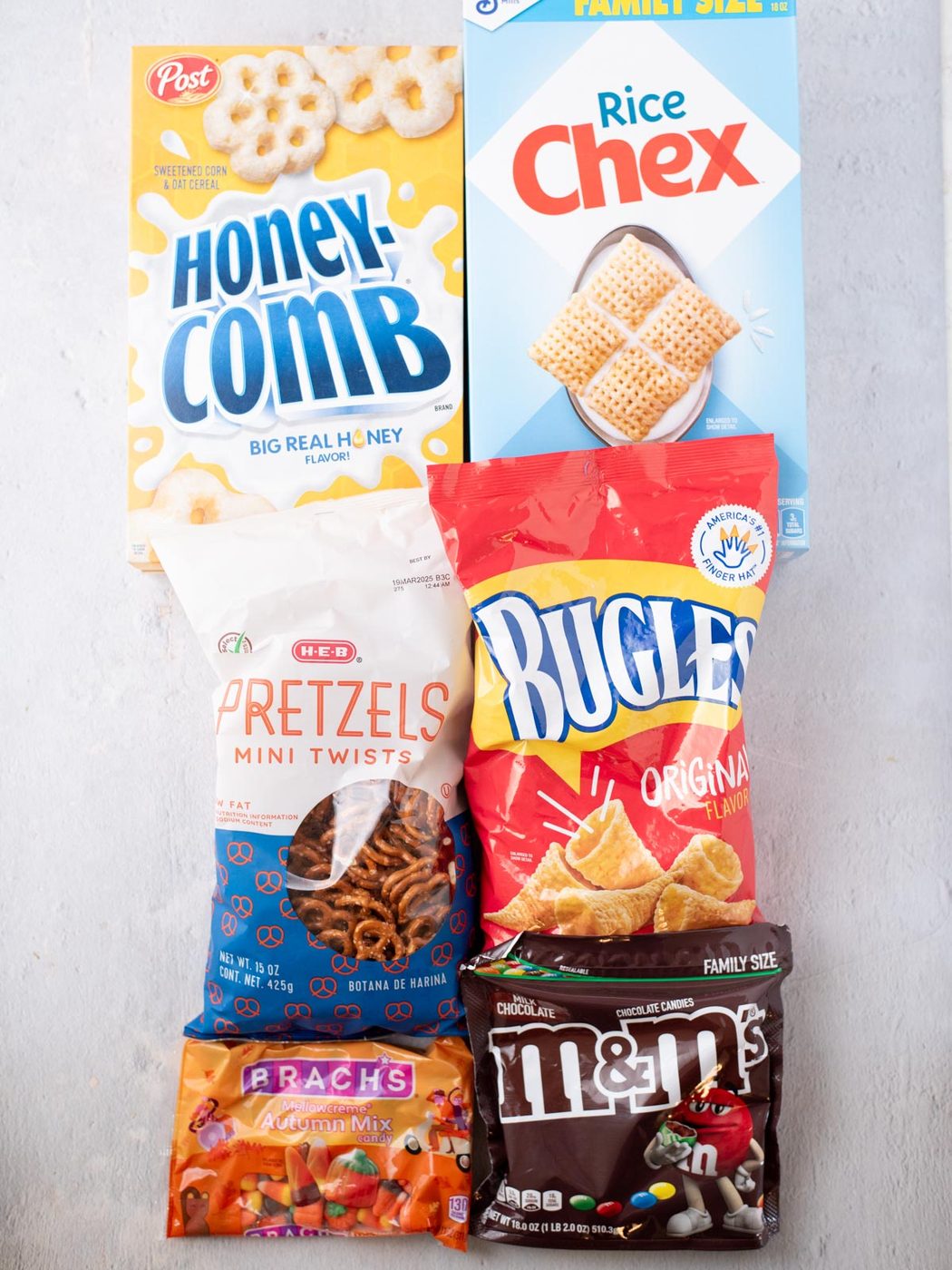 Six snack packages—Honeycomb cereal, Rice Chex, H-E-B pretzels, Bugles, Brach’s Autumn Mix candy, and M&M’s—arranged on a light surface in two rows.