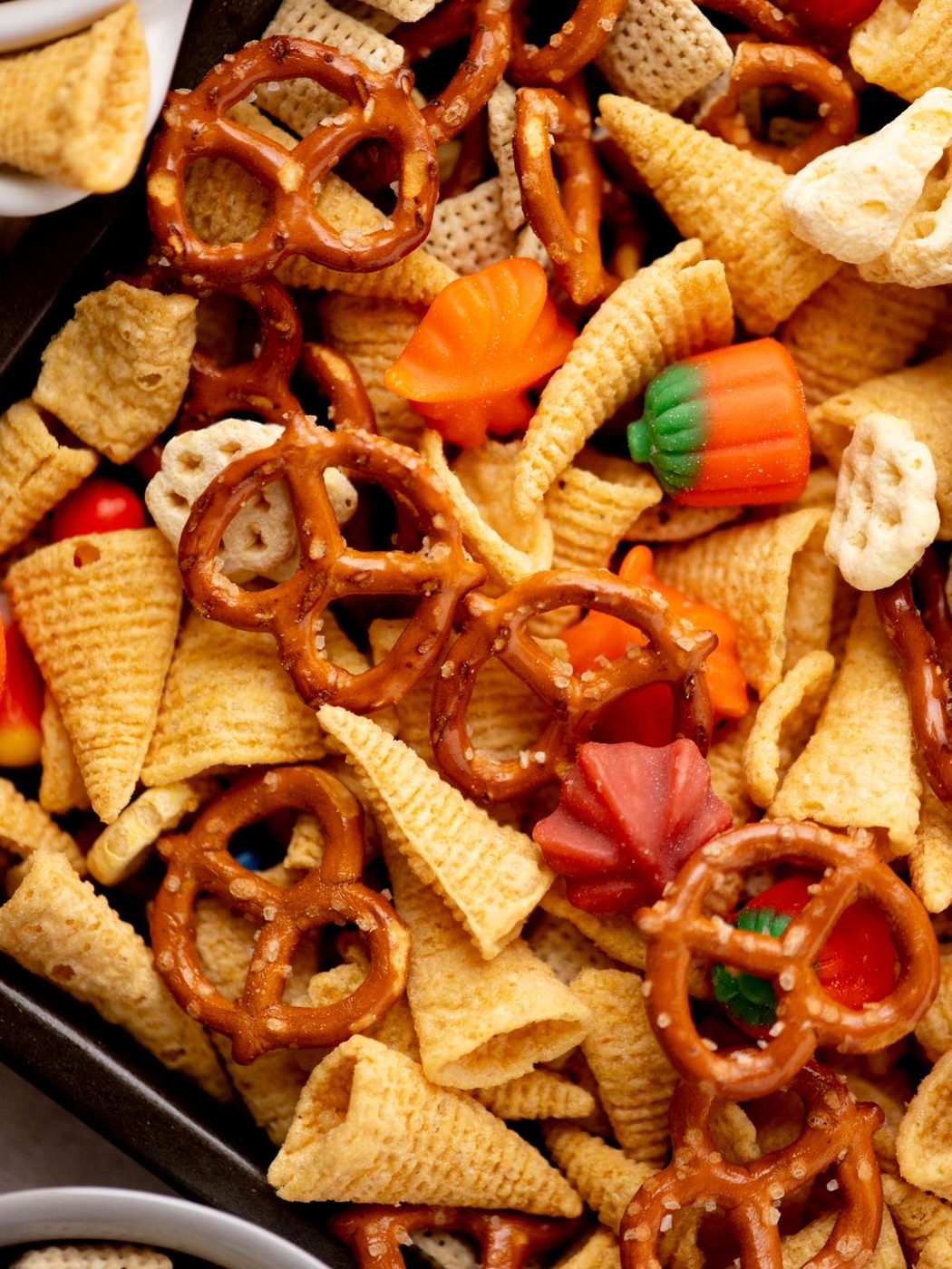 A close-up of a snack mix featuring pretzels, bugle chips, Chex cereal, and candy pumpkins and leaves in fall colors, all piled together in a tray.