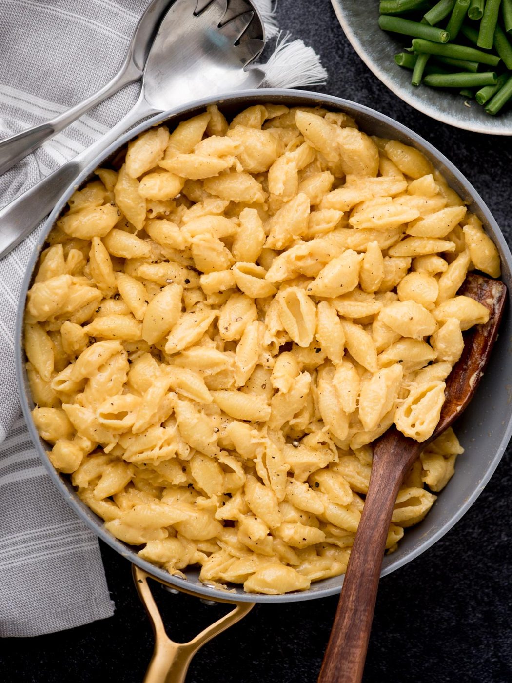 A skillet filled with creamy macaroni and cheese made from shell pasta, with a wooden spoon resting in it. A pair of serving utensils and a plate of green beans are beside the skillet on a dark surface.