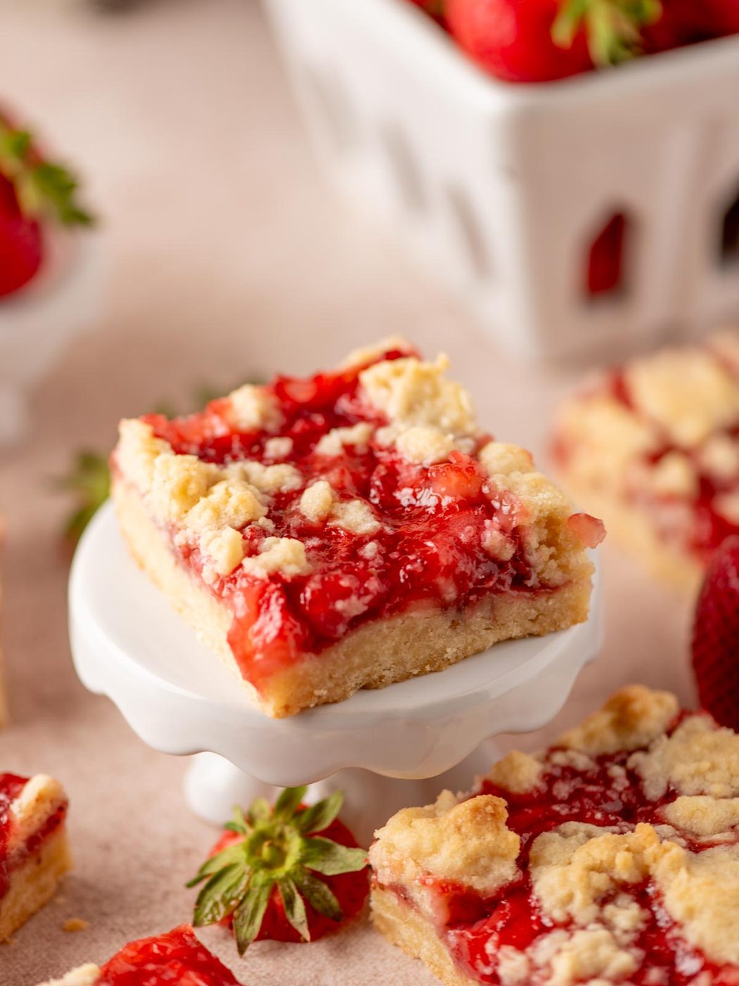 A close-up of a strawberry crumb bar on a small white stand. The bar has a buttery crust, a layer of vibrant red strawberry filling, and a golden crumb topping. Fresh strawberries and more bars are blurred in the background.