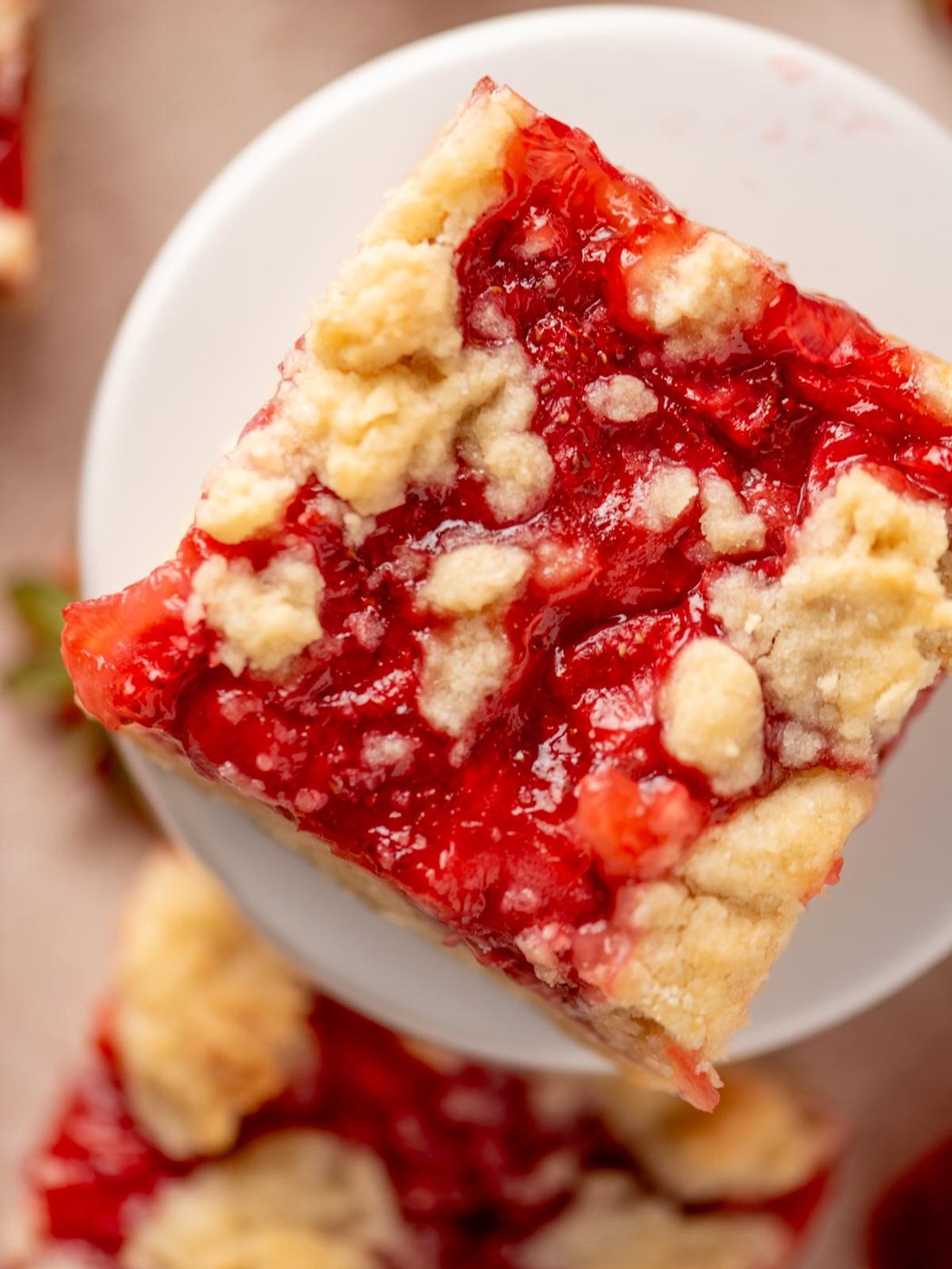 A close-up of a square strawberry crumble bar with a golden, crumbly topping and bright red strawberry filling, placed on a white plate.