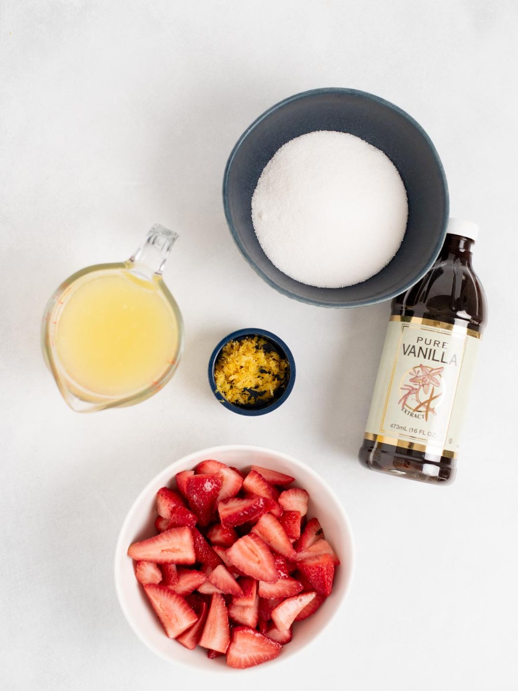 A bowl of sliced strawberries, lemon zest, lemon juice, sugar, and pure vanilla are arranged on a white surface—suited to making fresh strawberry lemonade at home.