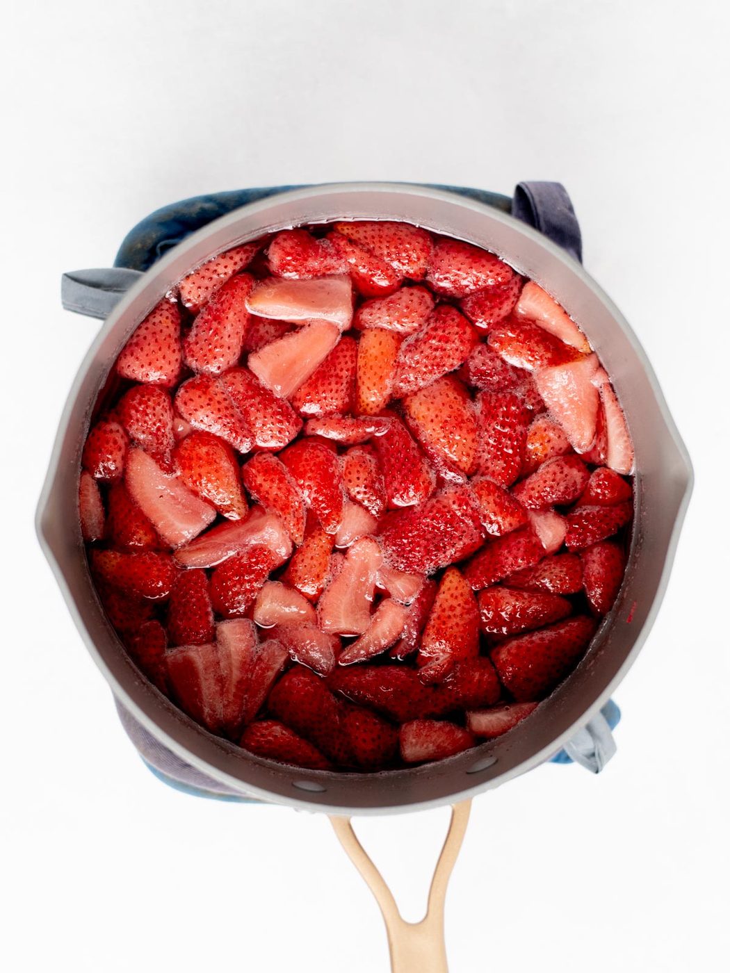 A pot filled with chopped strawberries sits on a blue oven mitt, viewed from above. The strawberries are bright red and appear to be cooking.