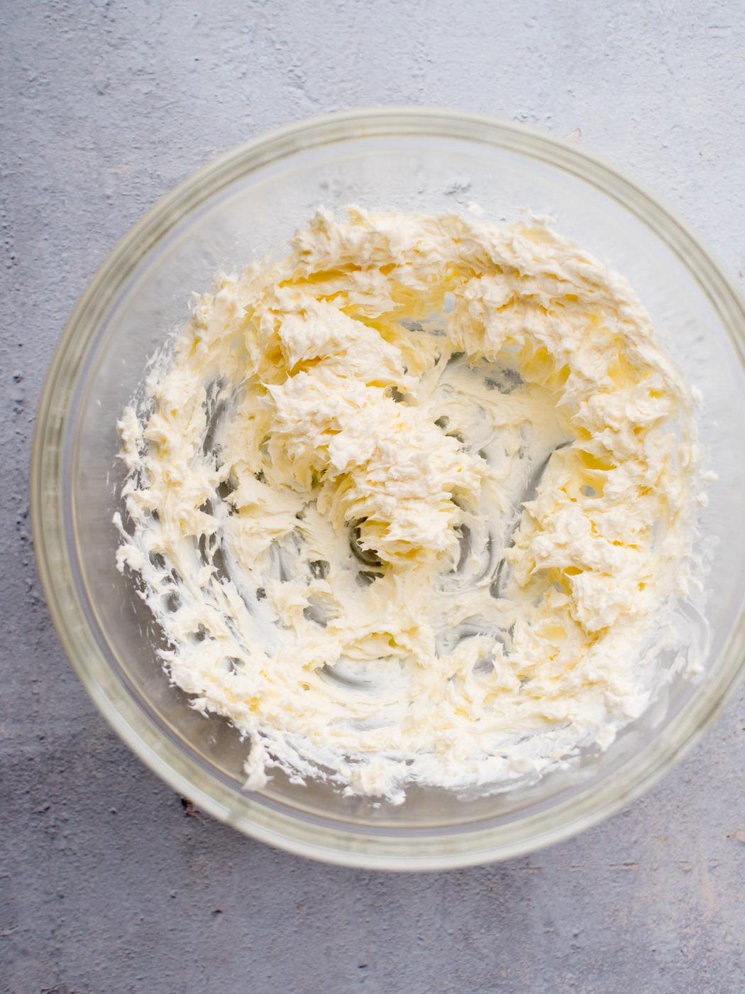 A clear glass bowl containing whipped cream cheese on a gray surface. The cream cheese appears fluffy and smooth, with swirls indicating it has been mixed thoroughly.