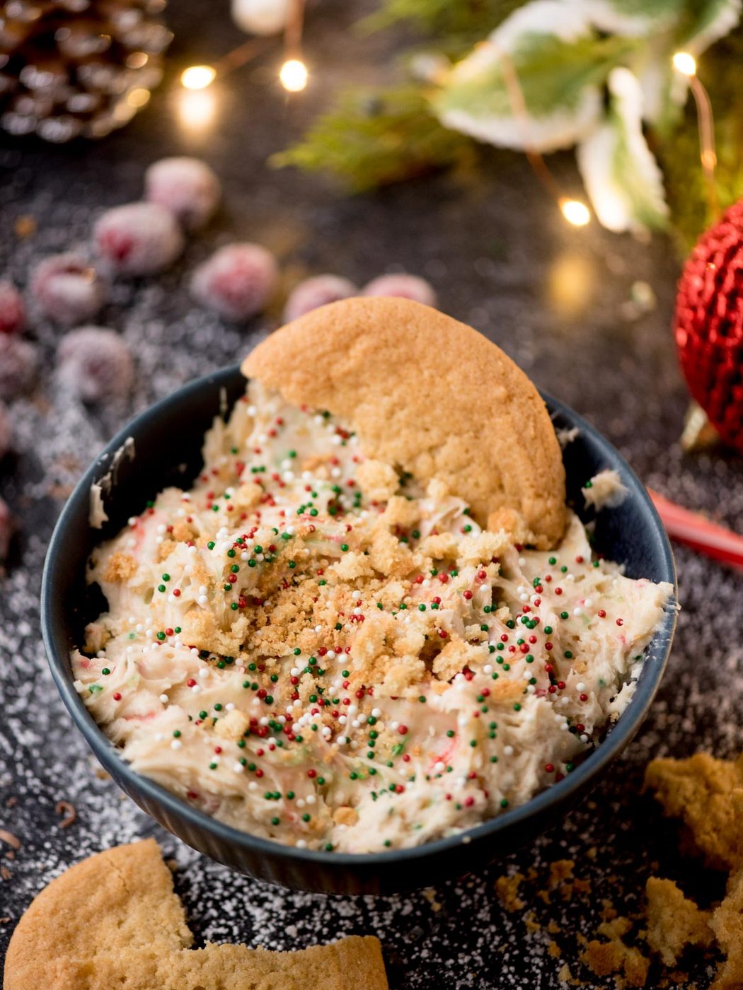 A bowl of creamy festive dip topped with red, green, and white sprinkles. A large cookie is placed in the dip. In the background, there are holiday decorations, frosted cranberries, and scattered cookie crumbs.