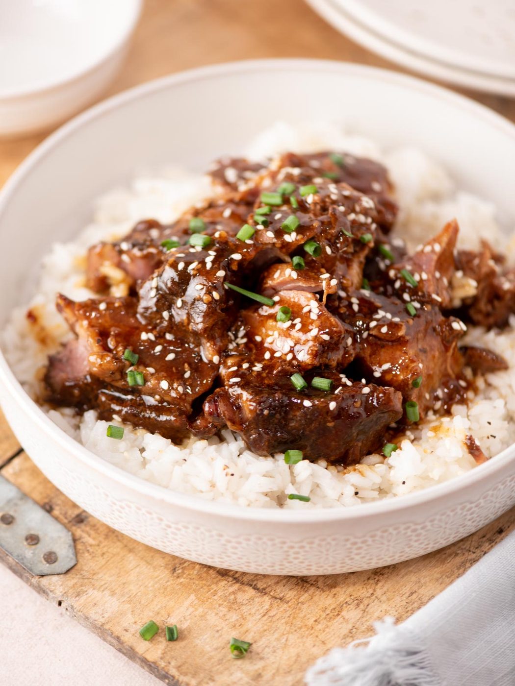 A bowl of white rice topped with glazed, saucy meat slices, sprinkled with sesame seeds and chopped green onions, on a wooden surface.