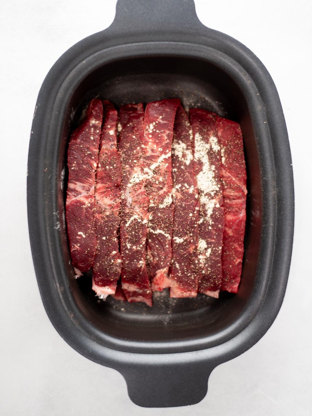 Raw beef slices with seasonings are arranged in a black slow cooker or roasting pan, ready for cooking. The photo is taken from above on a white background.