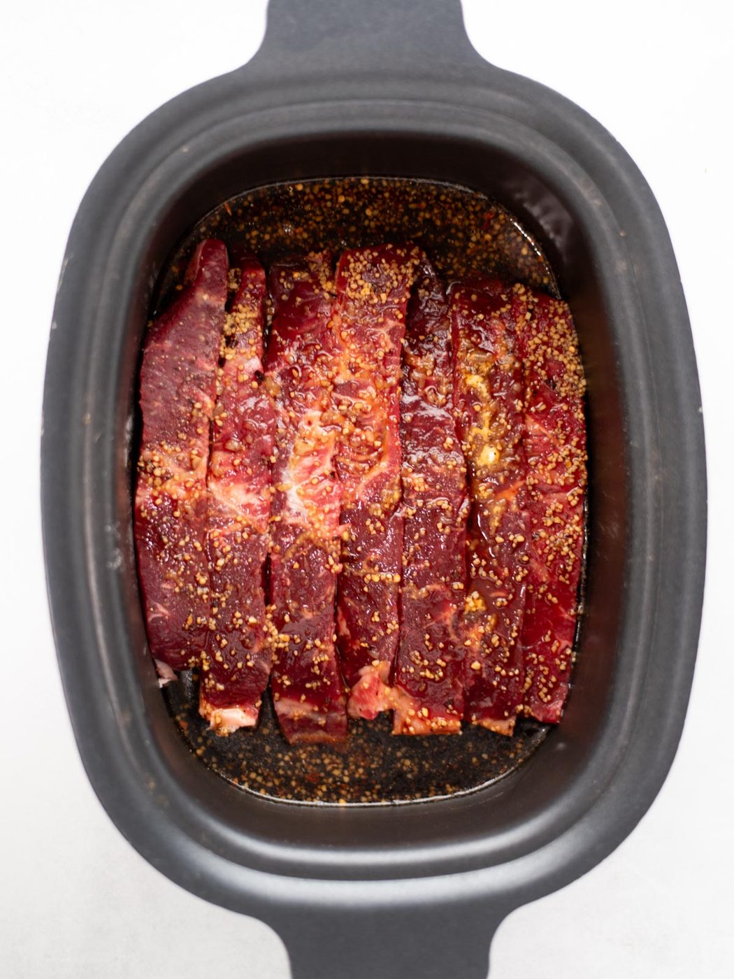 Slices of raw beef seasoned with spices are arranged in a black slow cooker, ready to be cooked.