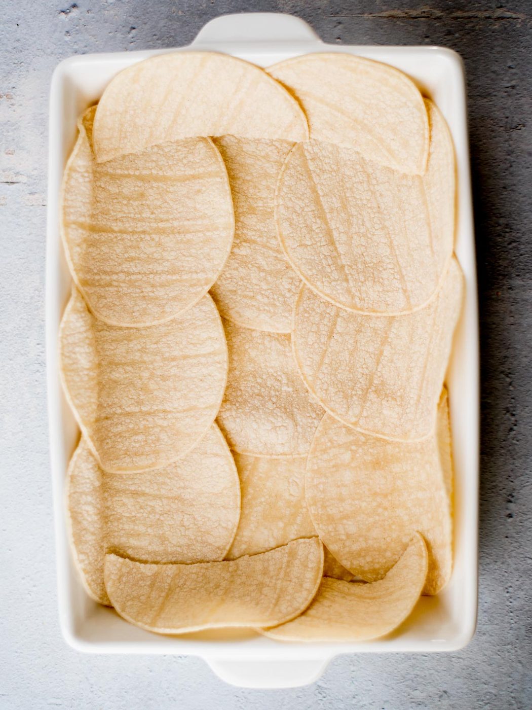 A rectangular baking dish filled with overlapping layers of round corn tortillas, placed on a textured gray surface.