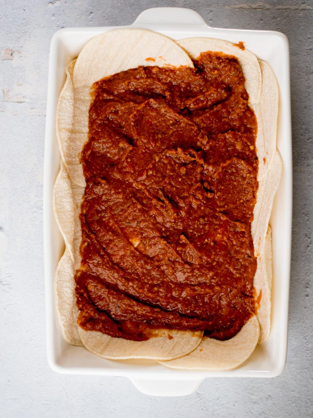 A rectangular baking dish filled with a layer of corn tortillas topped with red enchilada sauce, ready for further preparation. The edges of the tortillas are visible around the sauce. The dish is placed on a light textured surface.