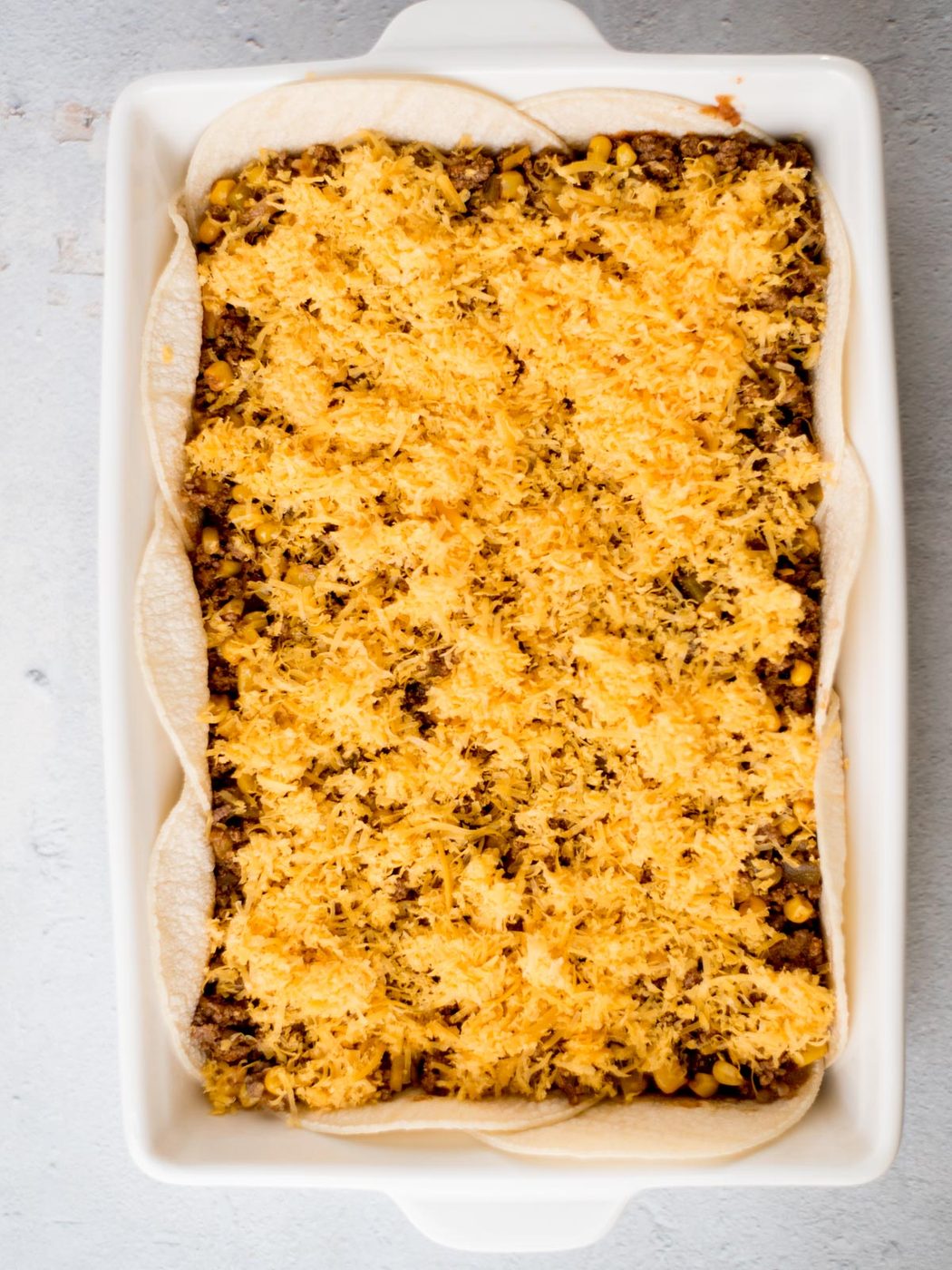 A rectangular baking dish filled with layers of tortillas, ground beef, corn, and melted cheese, creating a casserole. The dish is placed on a light gray surface.