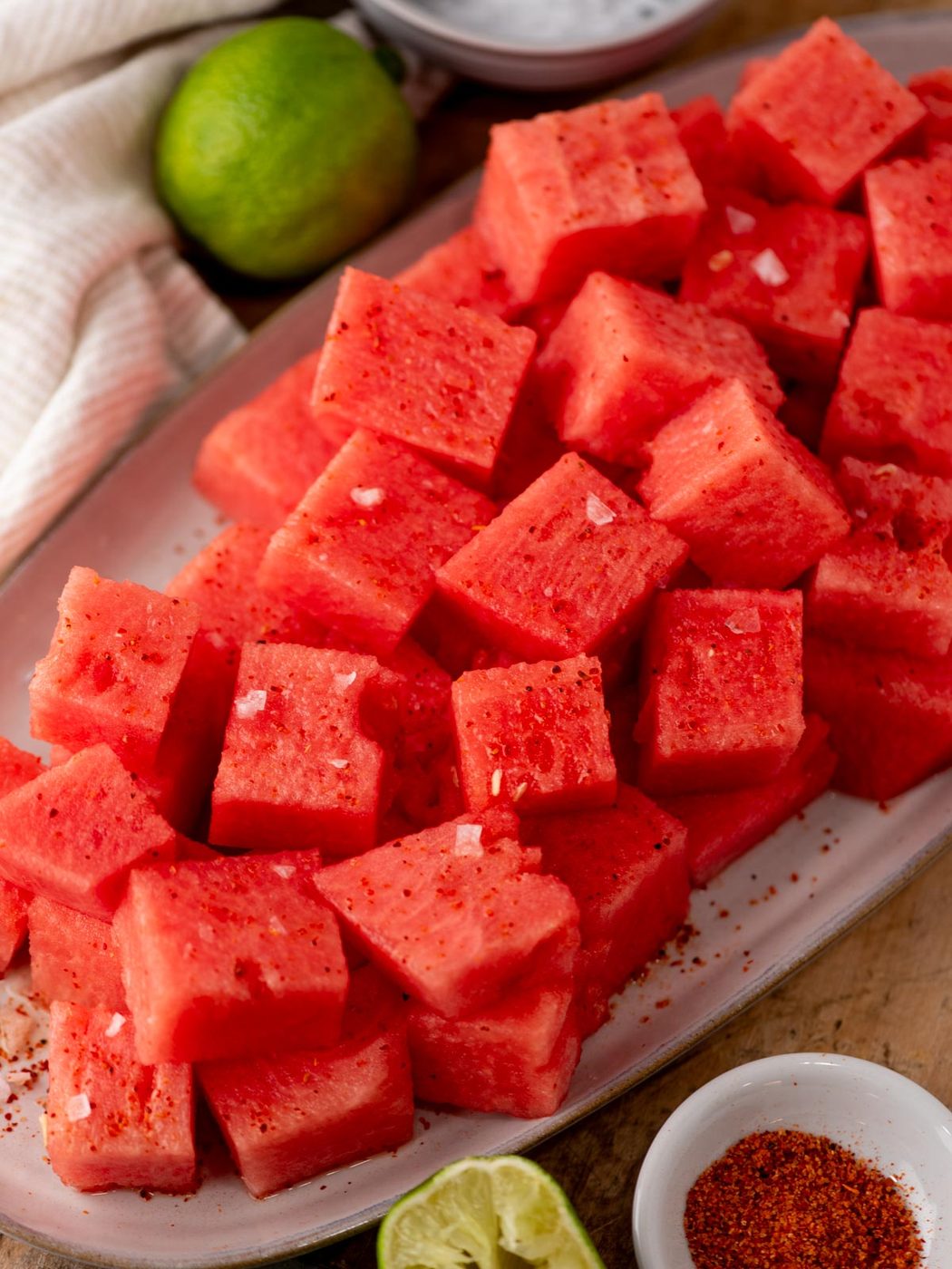 A platter of cubed watermelon sprinkled with chili powder and salt, surrounded by lime and chili seasoning bowls on a wooden surface.