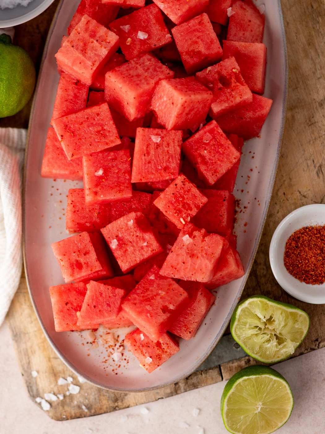 Cubed watermelon sprinkled with chili powder and coarse salt on a platter, surrounded by halved limes, a bowl of chili powder, and a wooden surface.