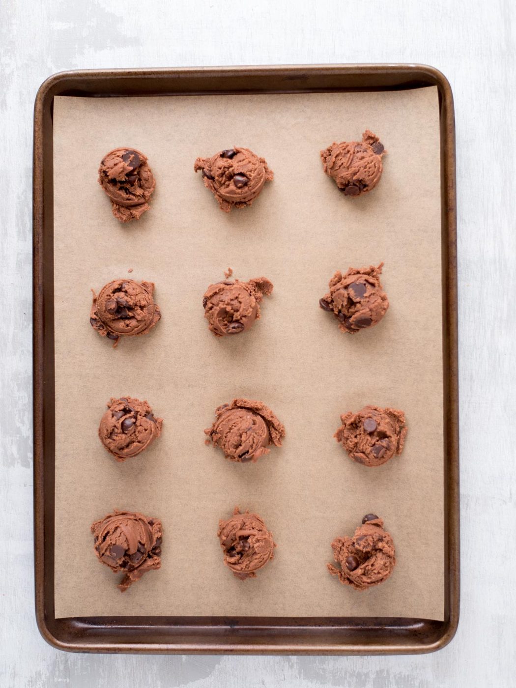 A baking tray lined with parchment paper holds twelve evenly spaced scoops of chocolate cookie dough, ready to be baked.
