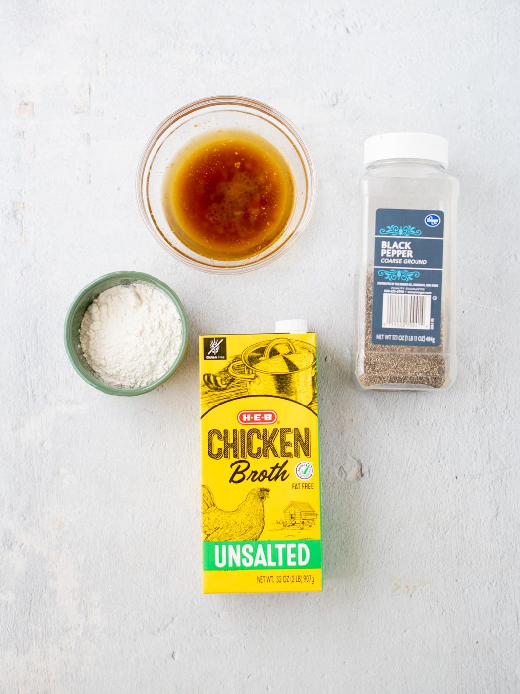 A bowl of brown liquid turkey gravy with drippings, a small bowl of white flour, a carton of unsalted chicken broth, and a container of ground black pepper are arranged on a light surface.
