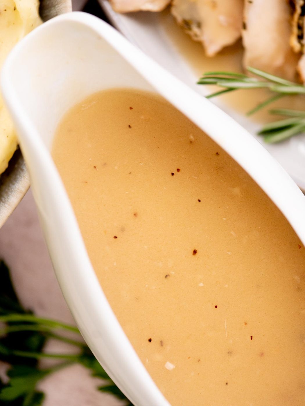 A close-up of a white gravy boat filled with creamy brown turkey gravy with drippings, speckled with pepper. Fresh herbs and a plate of sliced meat are slightly blurred in the background.