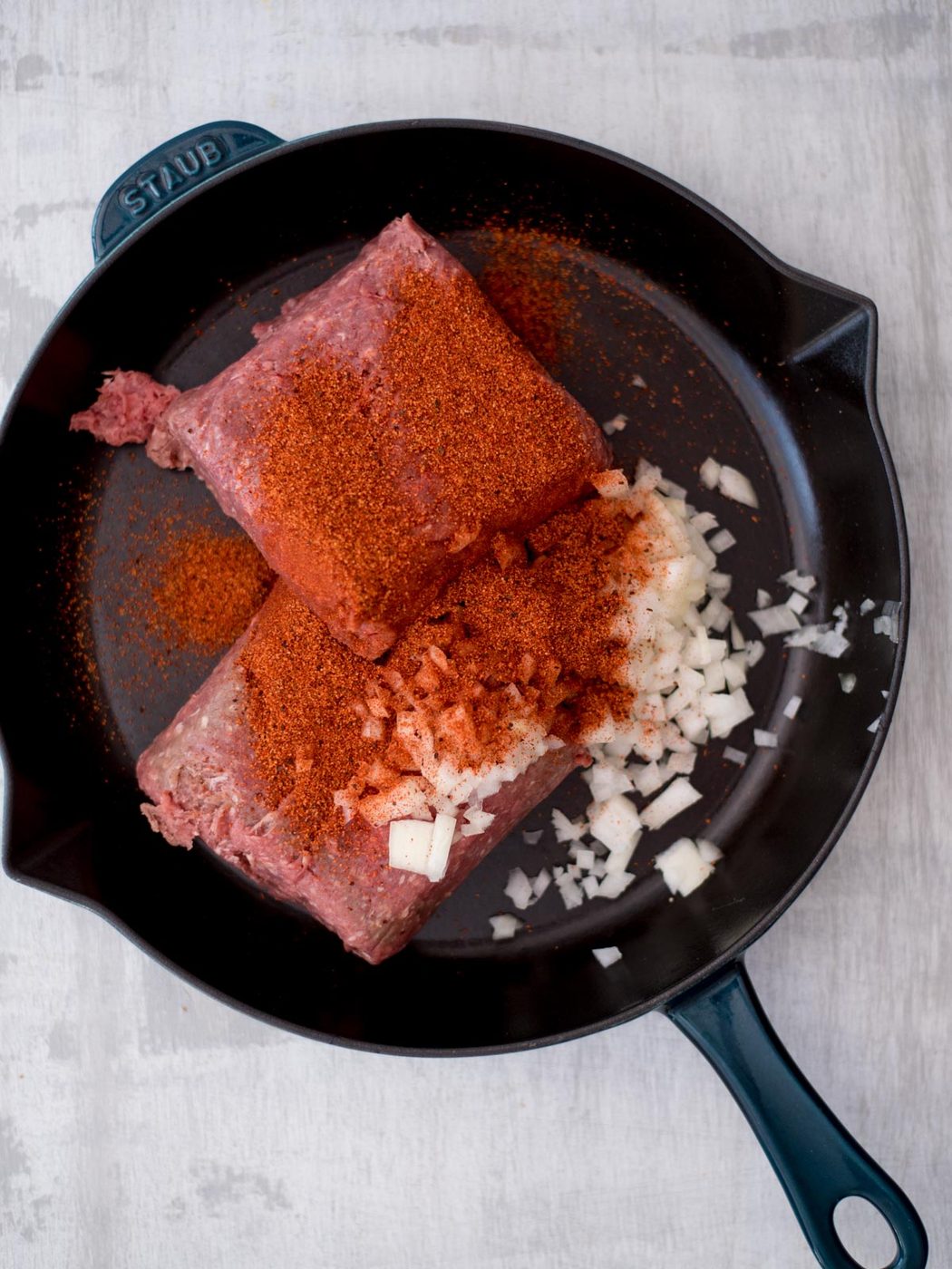 Raw ground beef, seasoning and diced onions added to a cast iron pan.