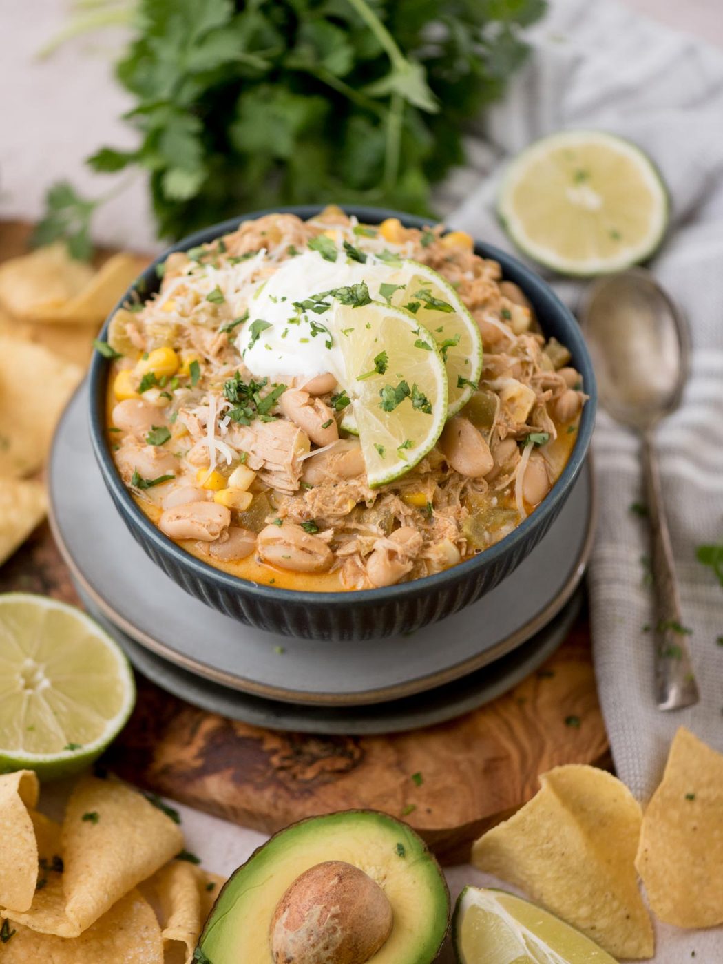 A bowl of white chicken chili garnished with lime slices, sour cream, and cilantro. Ingredients like avocado, tortilla chips, and lime halves surround the bowl. A spoon and a bunch of cilantro are placed nearby.
