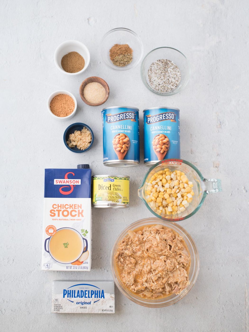 Ingredients for a southern white chicken chili are artfully arranged on the surface, featuring chicken stock, cream cheese, canned cannellini beans, diced green chiles, corn, shredded chicken, and several small bowls of spices and seasonings.