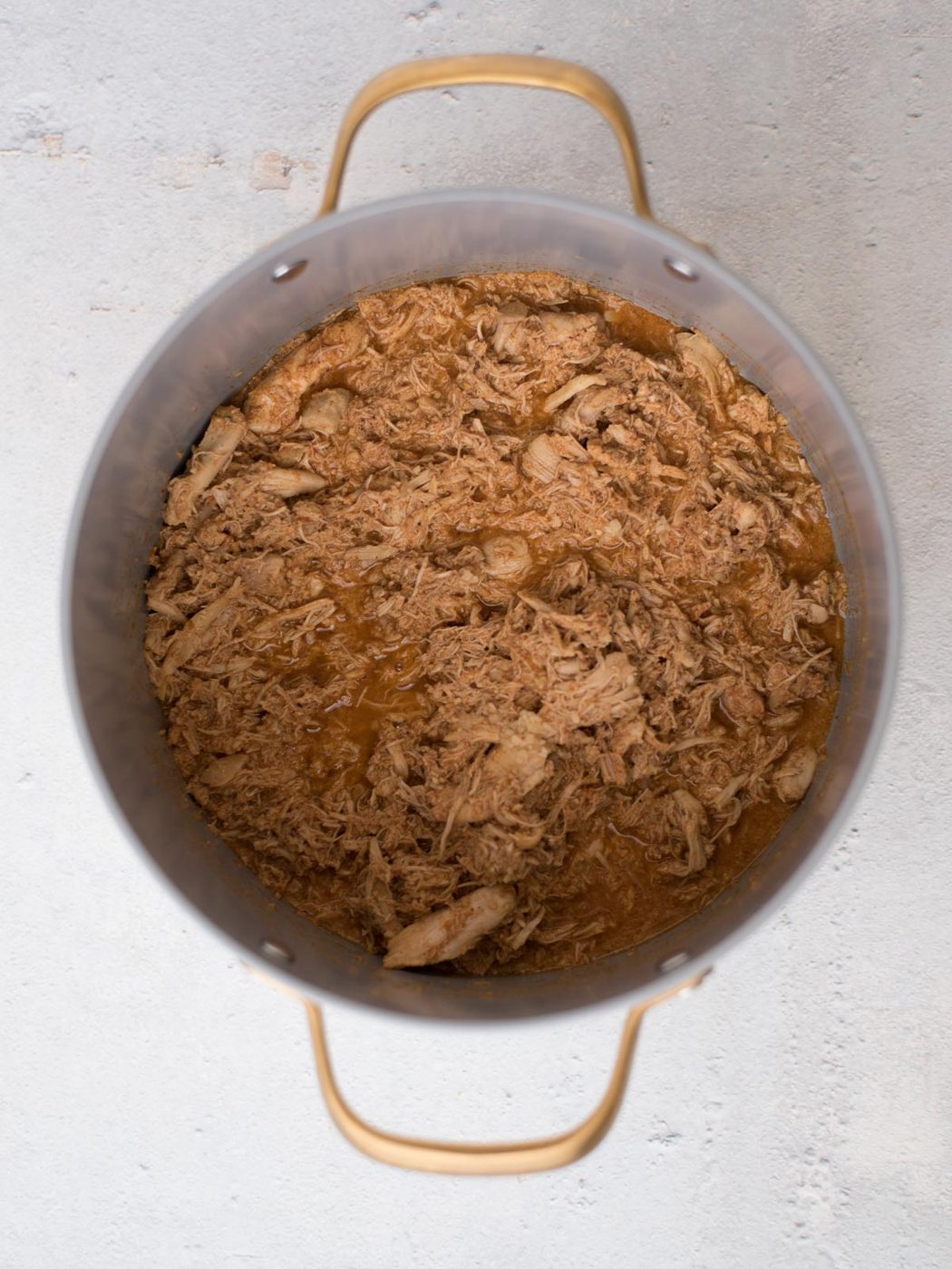 Top view of a large metal pot filled with tender, juicy shredded meat in a brown sauce for straightforward white chili with chicken. The pot, complete with handles on either side, rests on a light-colored surface.
