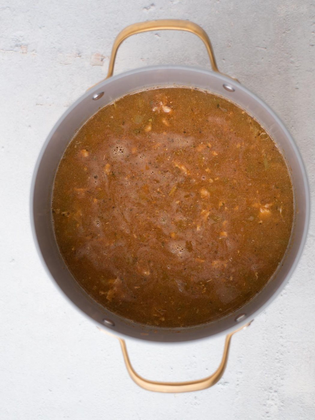 A pot filled with a savory white chicken chili, gently simmering to reveal tender pieces of meat and vibrant vegetables. The pot, equipped with two sturdy handles, sits gracefully on a light grey surface.