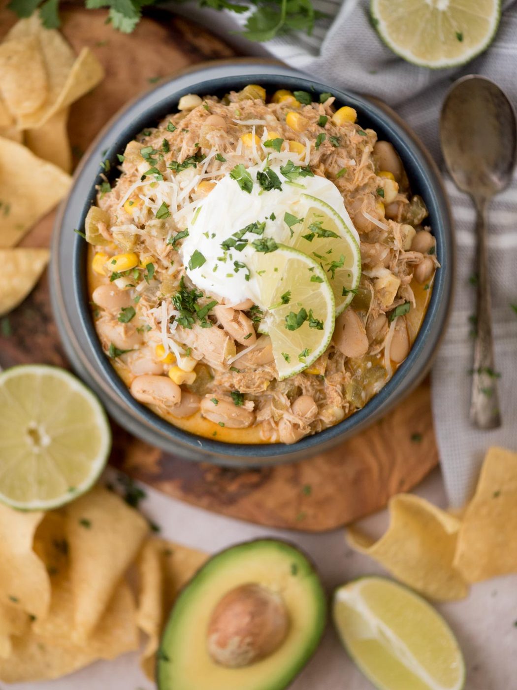 A bowl of white chicken chili garnished with sour cream, lime slices, and cilantro. Surrounded by avocado halves, lime, tortilla chips, and a spoon on a wooden board.