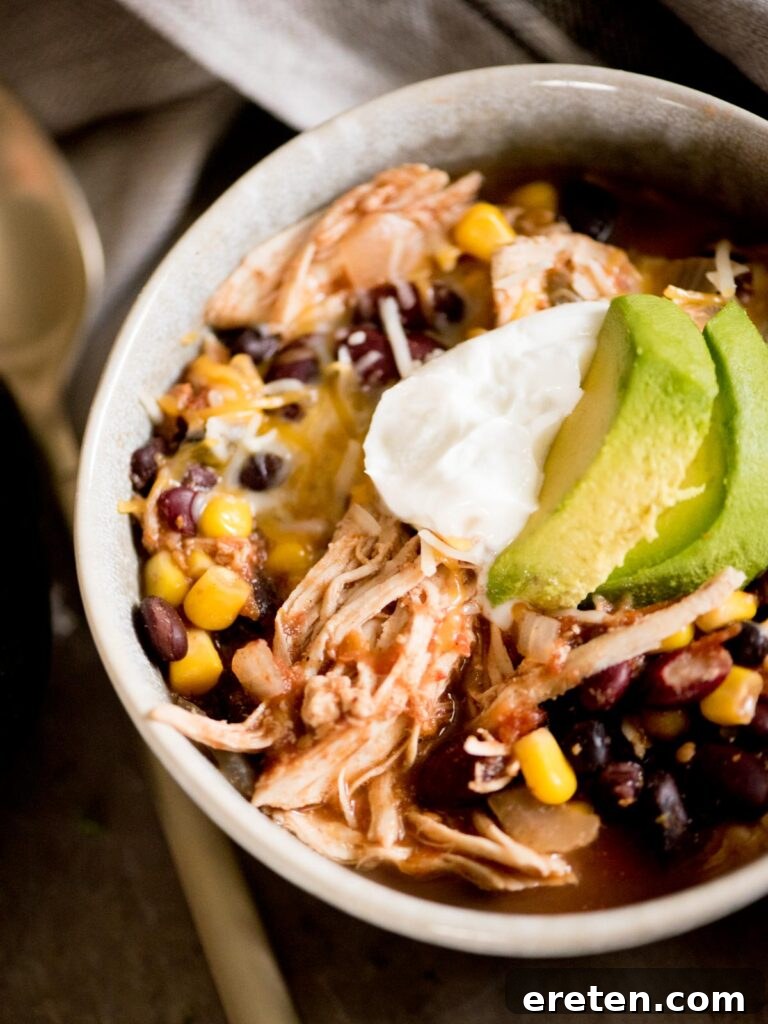 A close-up shot of Mexican chicken chili served in a rustic bowl, beautifully topped with fresh sour cream and slices of ripe avocado.