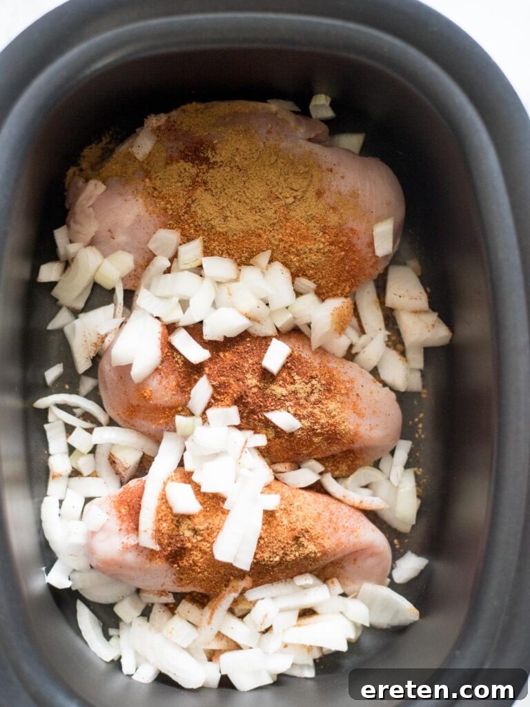 Finely chopped onions are added over the seasoned chicken in the slow cooker, ready to cook down and infuse flavor.