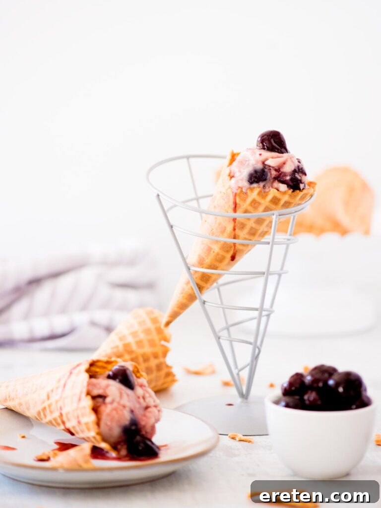 A scoop of homemade dark cherry ice cream in a waffle cone, with a bowl of fresh cherries and another ice cream cone on a plate.