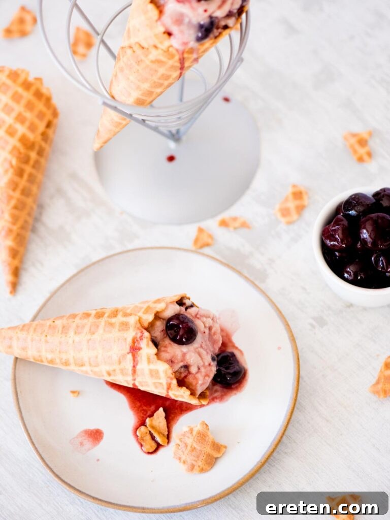 A scoop of black cherry ice cream in a waffle cone on a plate, surrounded by a bowl of fresh cherries and pieces of waffle cone.