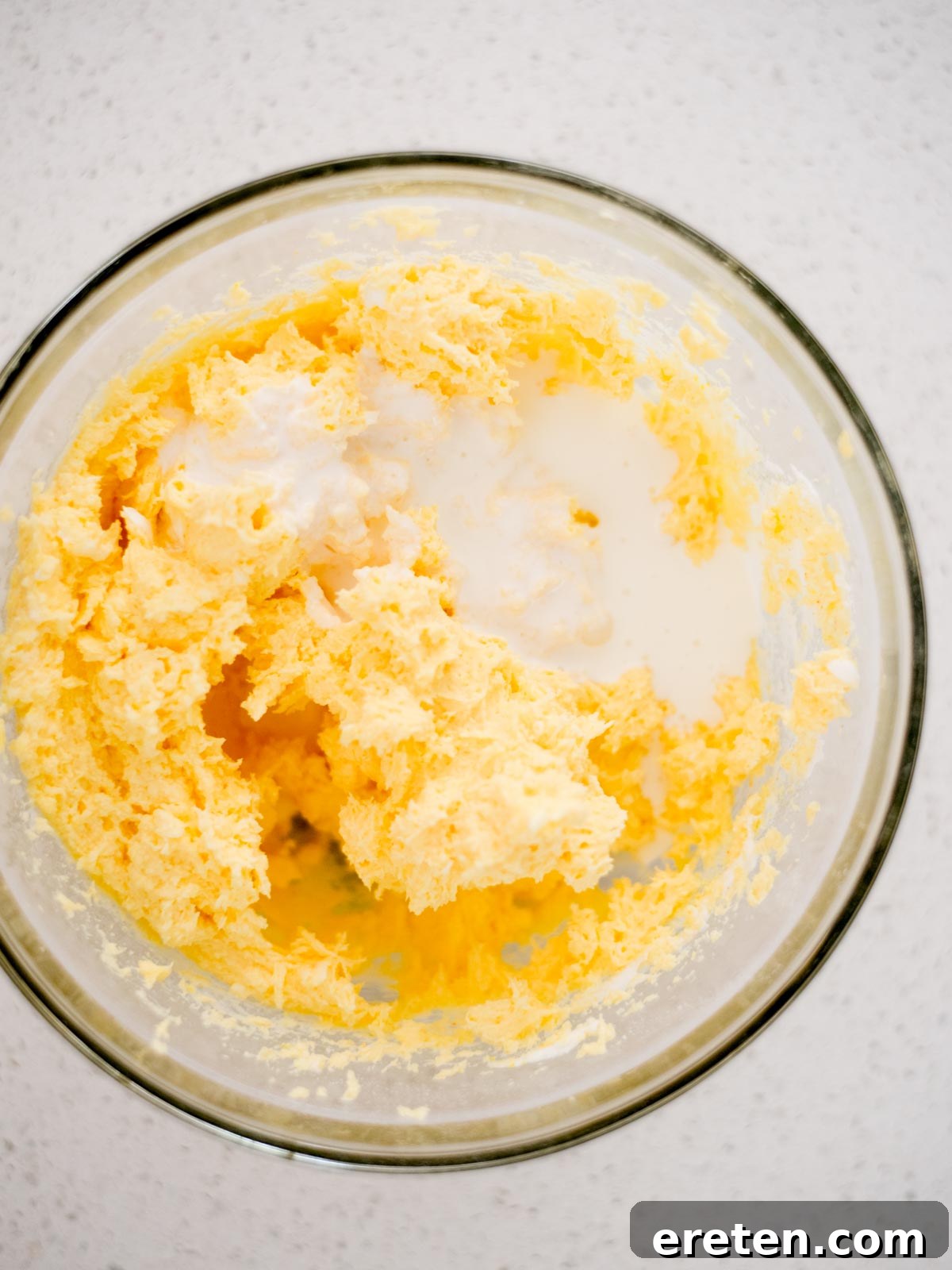 A close-up view showing whipping cream being added to a large mixing bowl already containing vanilla yogurt, cream cheese, and pudding mix, illustrating one of the key steps in preparing the fluffy fruit dip.