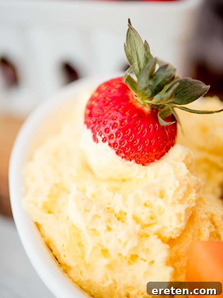 A single fresh strawberry, perfectly coated in a generous layer of creamy yogurt fruit dip, is held aloft, ready to be enjoyed. In the soft-focus background, a bowl of the delicious dip and a variety of colorful fruits await.
