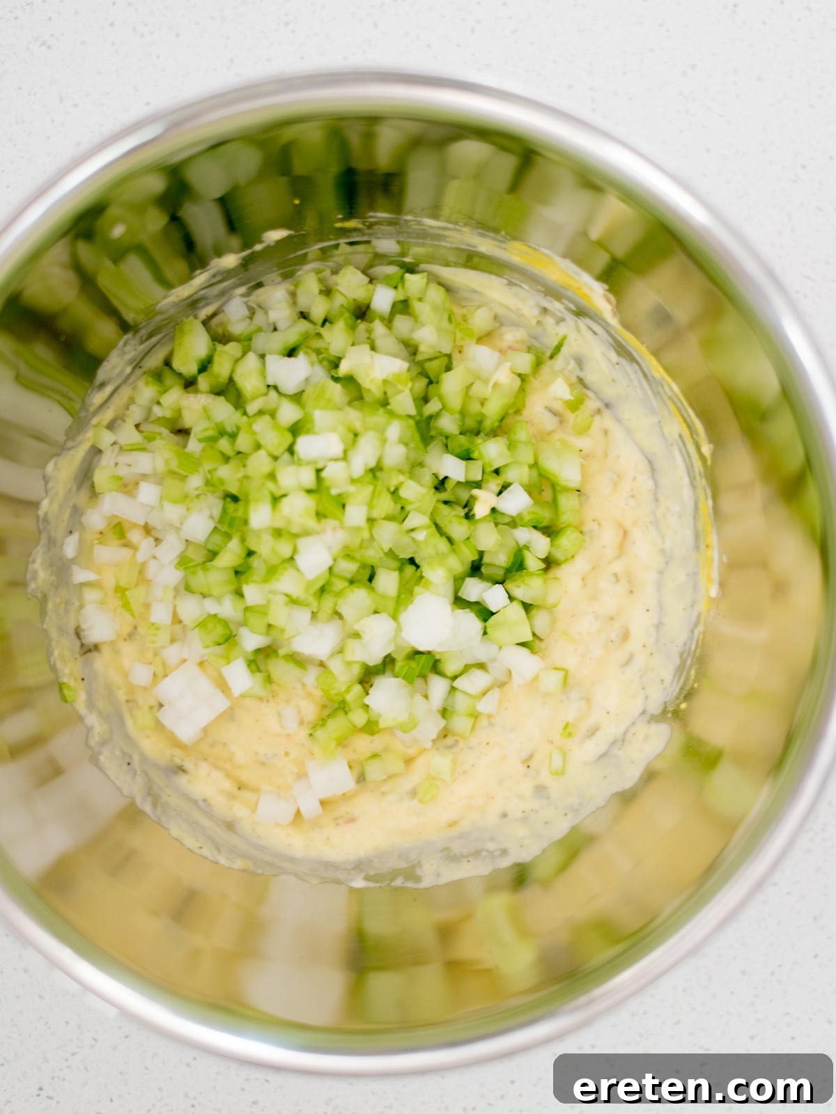 celery and onion added to potato salad dressing in a mixing bowl