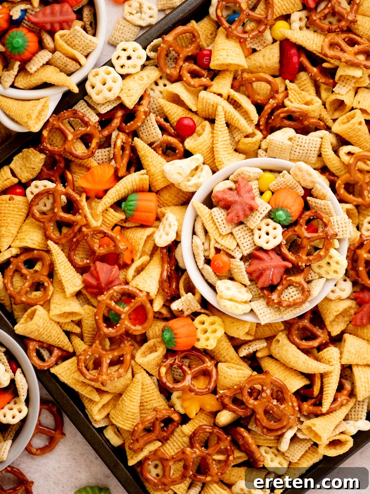 A festive snack mix featuring pretzels, Bugles, Chex cereal, candy pumpkins, and colorful candy-coated chocolates, displayed in bowls and scattered on a tray.