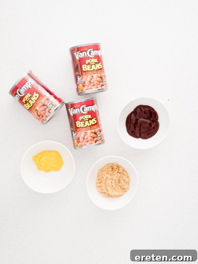 The four simple ingredients required to make smoked baked beans: cans of pork and beans, a bottle of barbecue sauce, mustard, and brown sugar, laid out on a wooden surface.