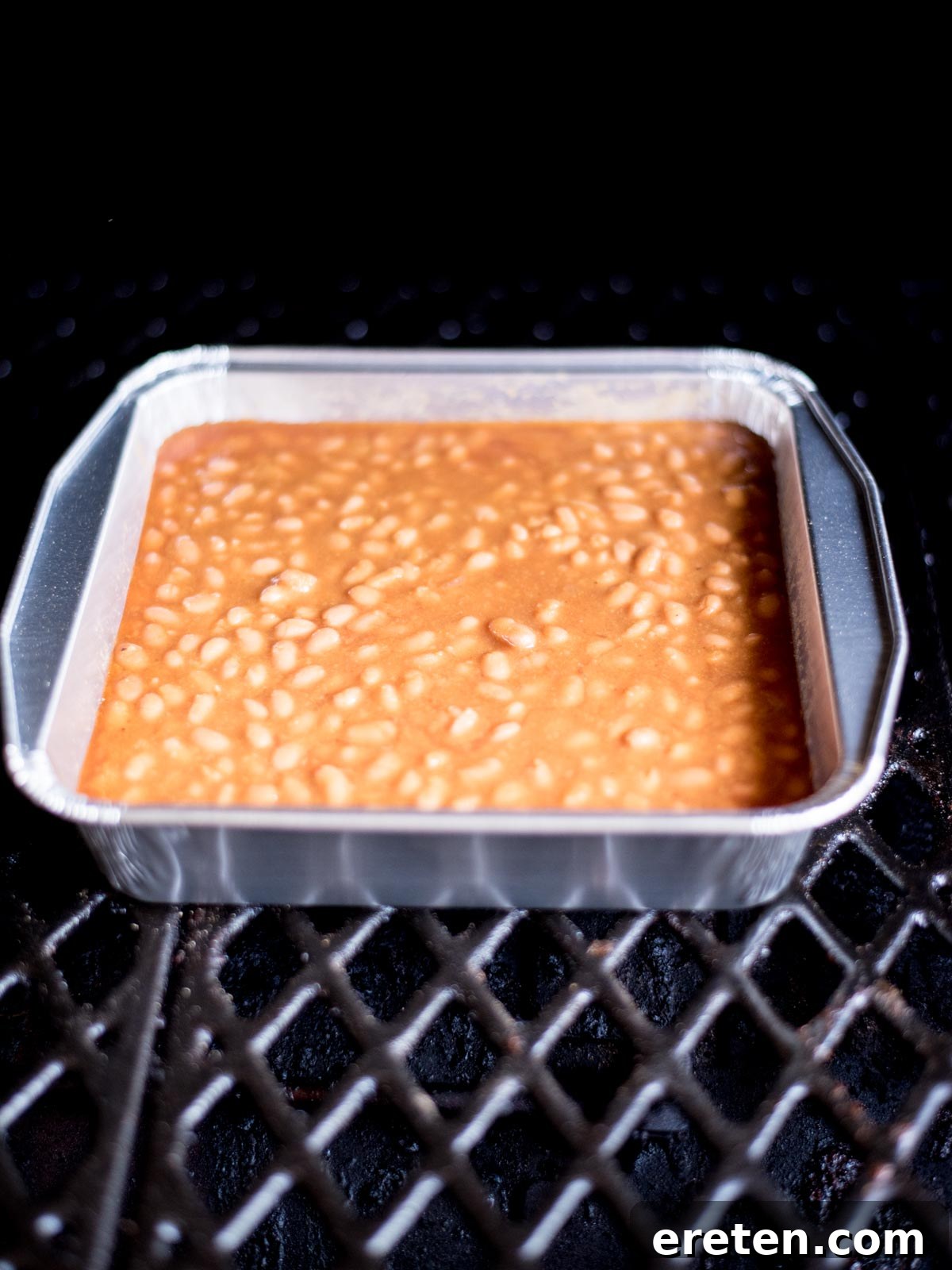 A close-up view of baked beans simmering in an aluminum pan inside a pellet smoker, surrounded by a gentle haze of smoke.