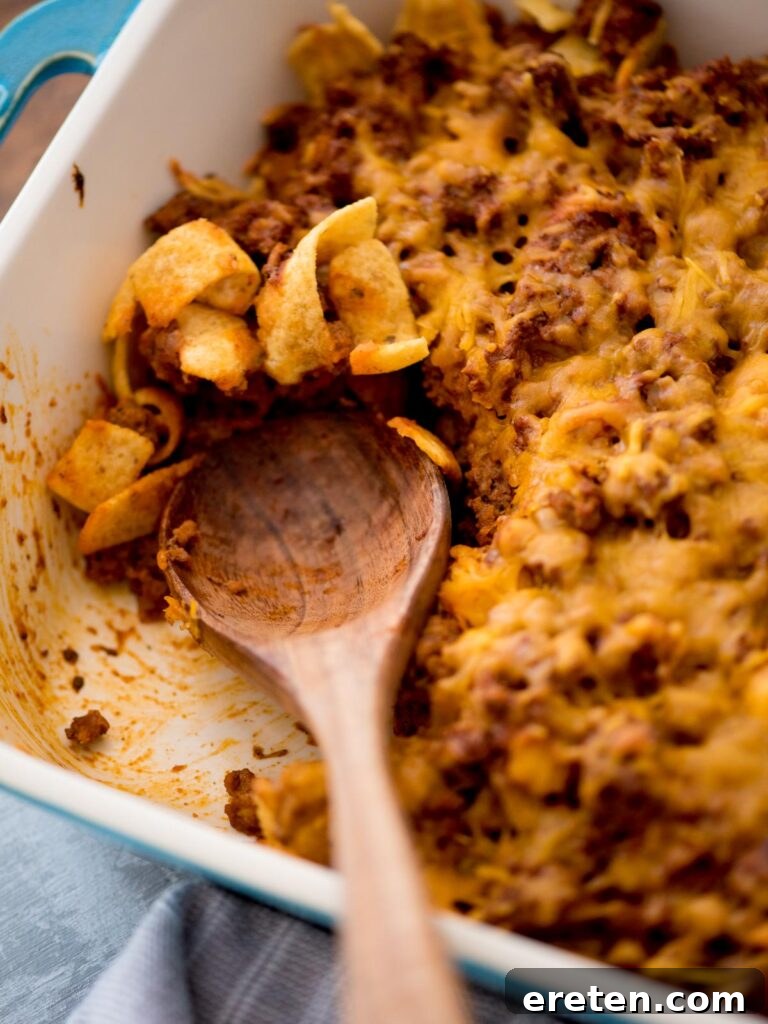 A wooden serving spoon rests in a casserole dish of Walking Taco Casserole, with a portion already served, highlighting the delicious layers.
